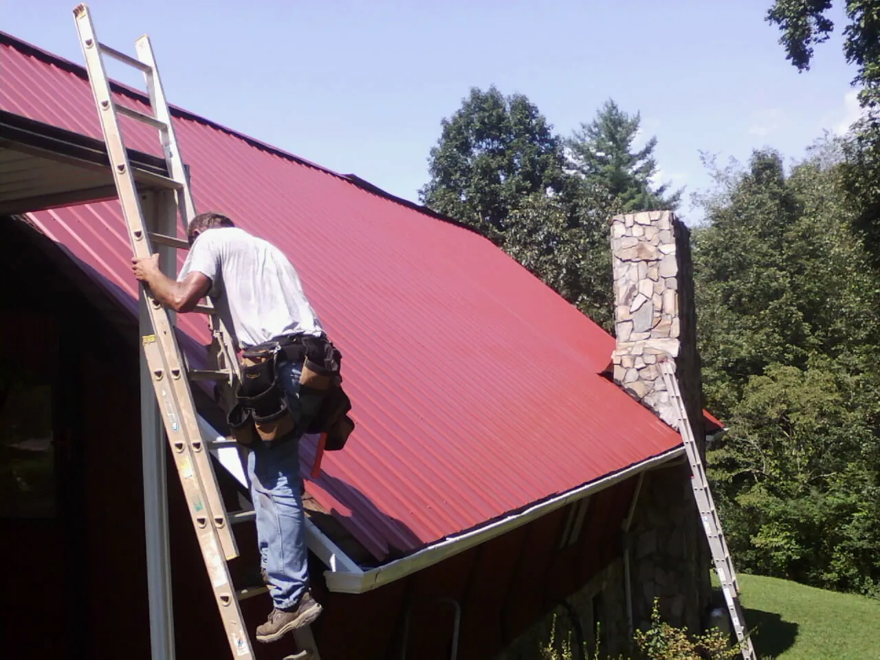 Roofer on a ladder beside a red metal roof and stone chimney — Soffit & Fascia Repair by Roofing Brothers in Bee Spring